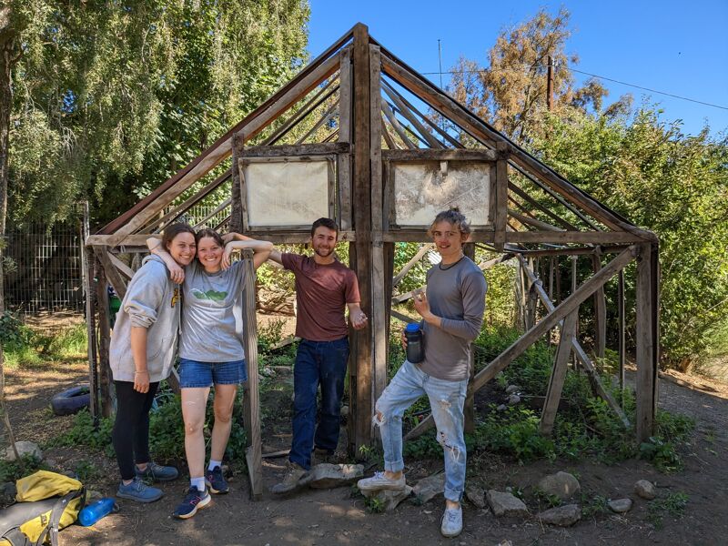 The image shows four people standing in front of a wooden structure that resembles a greenhouse frame. The structure is open, with some translucent panels in the upper sections. The people are smiling and appear to be posing for the photo. The setting is outdoors, with trees and greenery visible in the background. The sky is blue with some clouds.